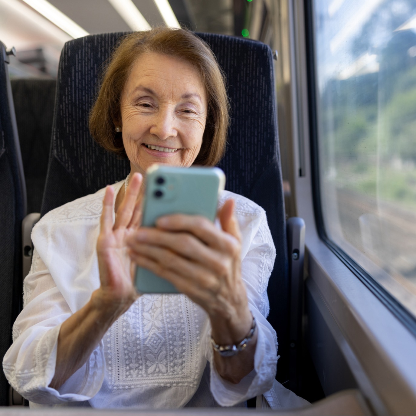An older woman smiles while using a smartphone on a train, wearing a white blouse, with greenery visible through the window.
