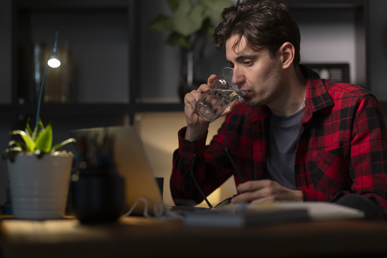 A young man in a red plaid shirt drinks water at a dimly lit desk with a laptop and small plant nearby.