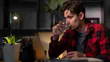 A young man in a red plaid shirt drinks water at a dimly lit desk with a laptop and small plant nearby.