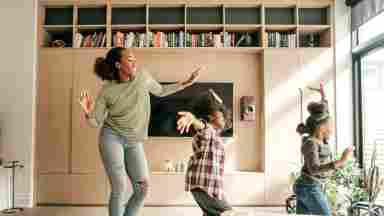 A Black mother and her two children dance joyfully in the living room together.