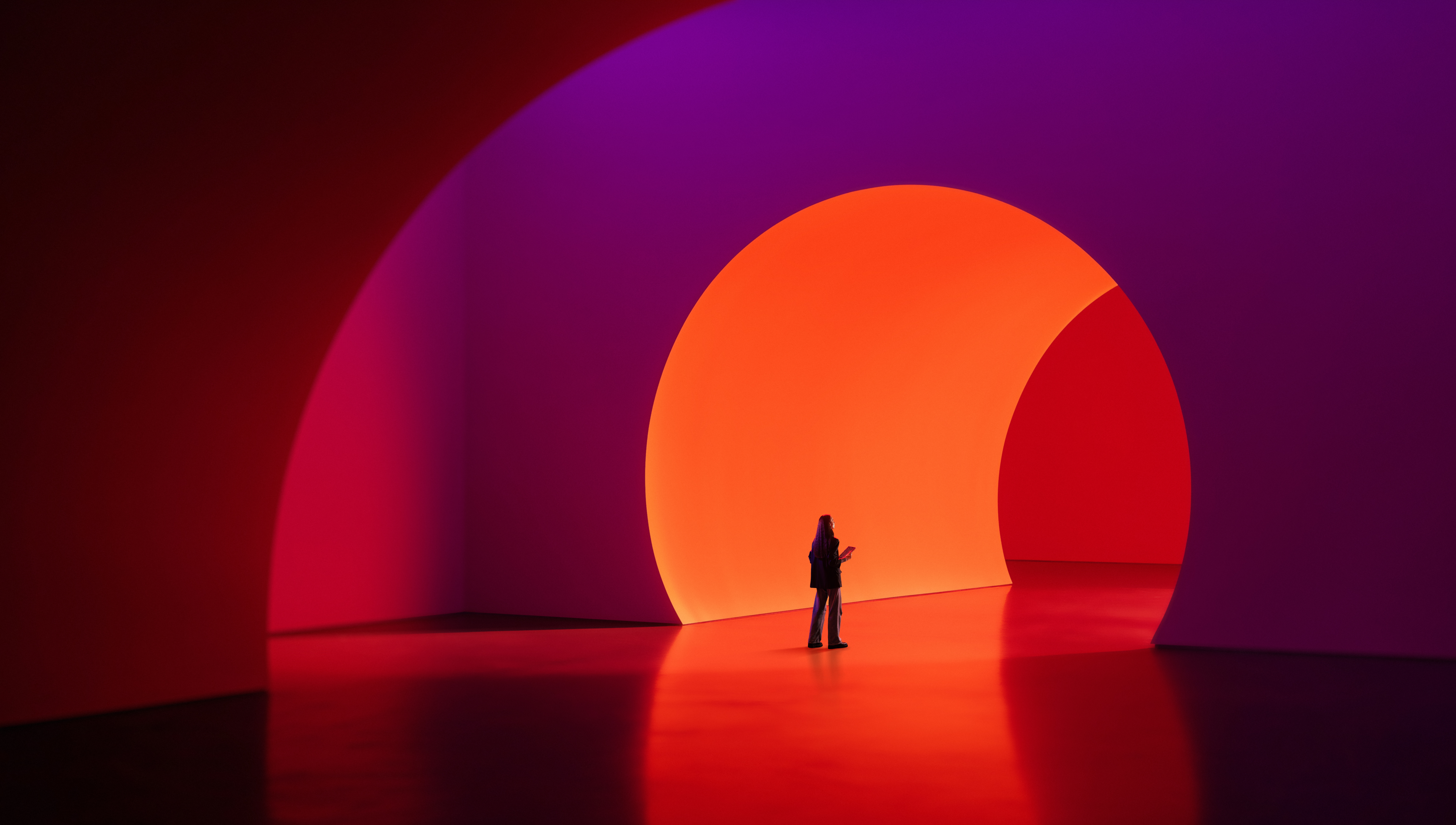 A lone person stands before a large glowing orange circular arch in a vibrant red and purple immersive art installation.