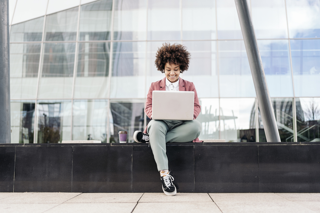 Businesswoman with afro hair wearing formal clothes sitting outdoors in business area and using her computer.