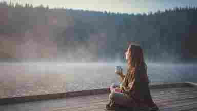 Woman relaxing on a pier by by a calm, natural lake in the early morning. She is holding a steaming mug.