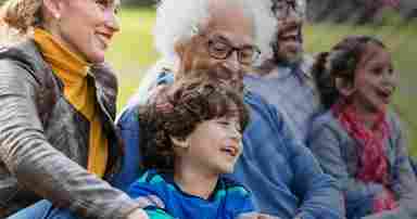 Portrait of a multi-generation family, sitting together on the grass in a park. The main focus is the 6 year old boy laughing in the foreground next to his mother and grandfather. The grandfather is in his 80s.