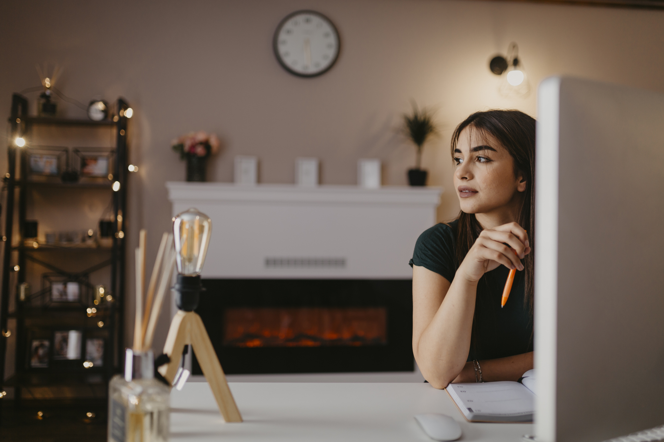 Person in dark shirt sits at computer in cozy room with fireplace, bookshelf and wall clock, taking a moment to disengage from online cognitive training and look out the window,