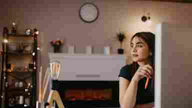 Person in dark shirt sits at computer in cozy room with fireplace, bookshelf and wall clock, taking a moment to disengage from online cognitive training and look out the window,