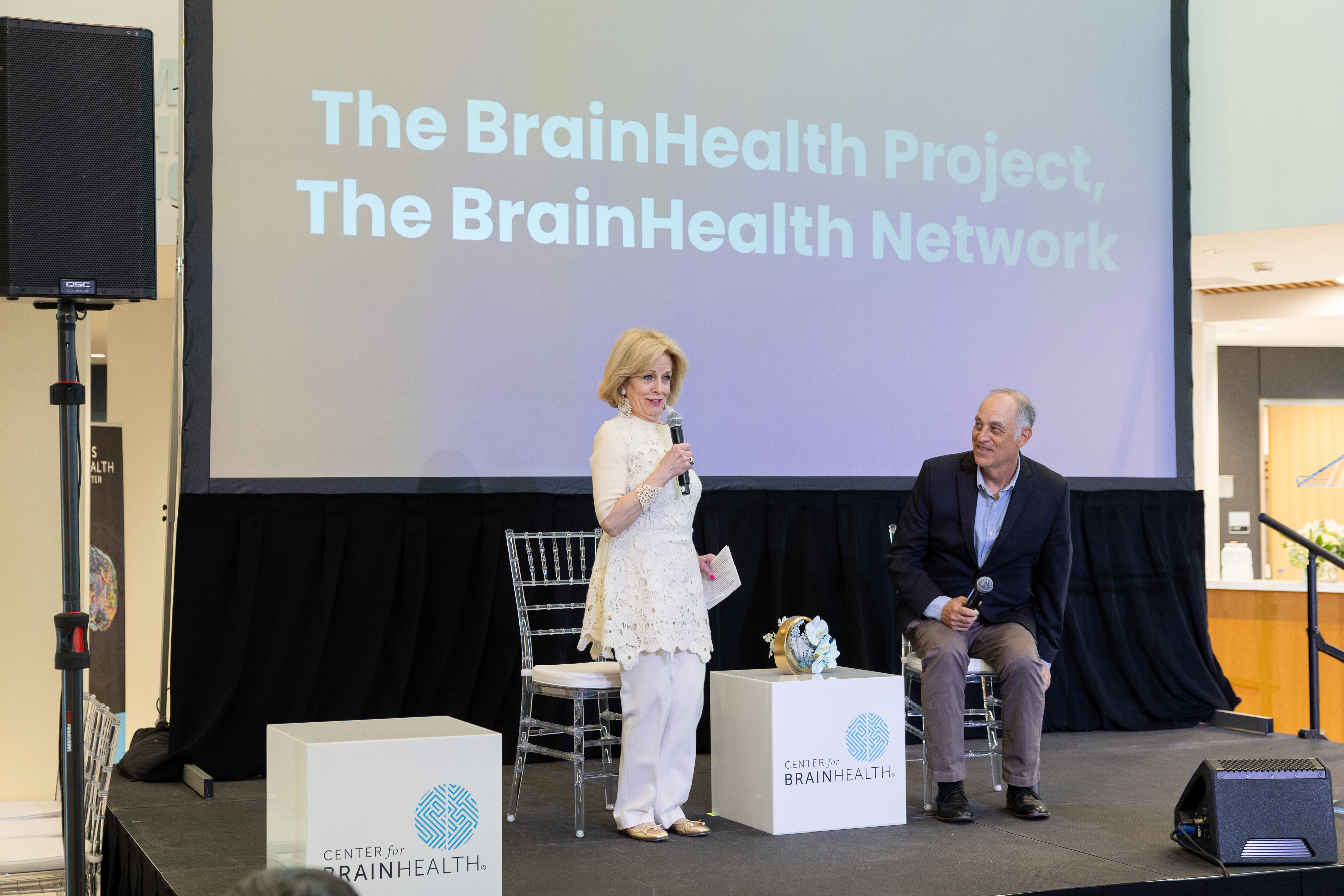 Dr. Sandi Chapman in white speaking while Dr. Mark D' Esposito in dark suit listens on stage at BrainHealth Week 2026 Science Summit. Screen behind reads The BrainHealth Project, The BrainHealth Network. 