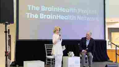 Dr. Sandi Chapman in white speaking while Dr. Mark D' Esposito in dark suit listens on stage at BrainHealth Week 2026 Science Summit. Screen behind reads The BrainHealth Project, The BrainHealth Network.