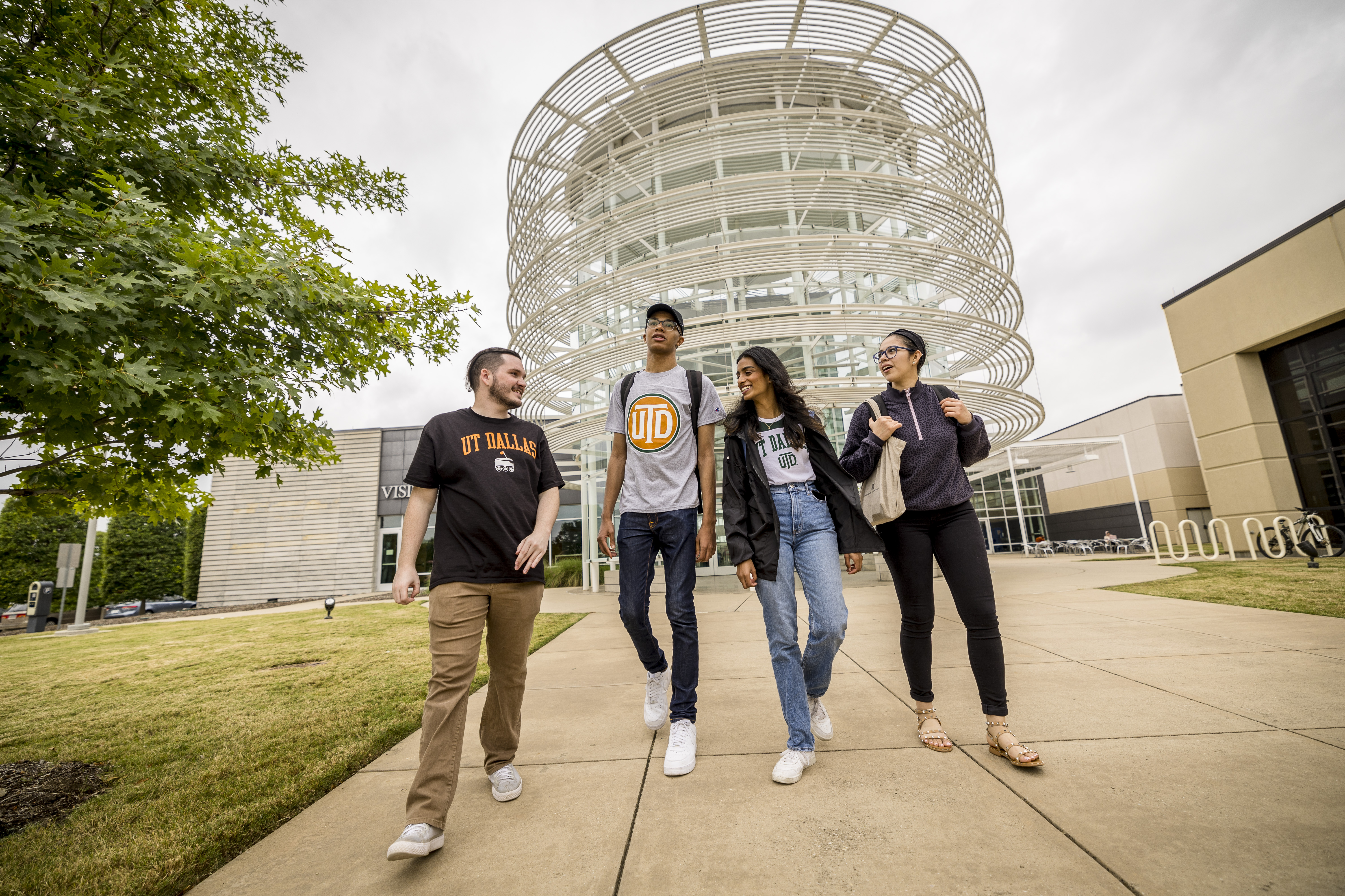 UT Dallas students walk briskly while in conversation outside the modern architecture of the UT Dallas' Activity Center.