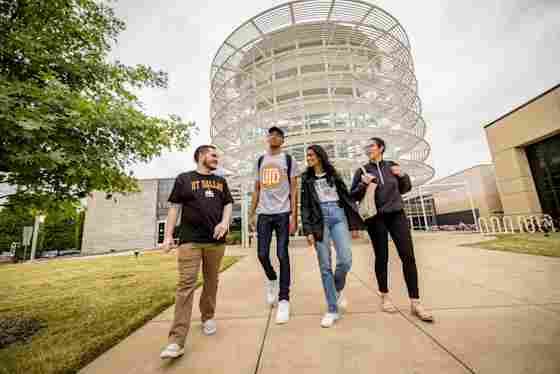 UT Dallas students walk briskly while in conversation outside the modern architecture of the UT Dallas' Activity Center.