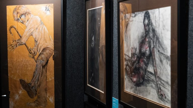 Three framed artworks displayed on dark gallery walls, featuring expressive figure drawings in warm amber and dark charcoal tones.