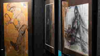 Three framed artworks displayed on dark gallery walls, featuring expressive figure drawings in warm amber and dark charcoal tones.