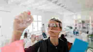 A young woman writing on a transparent wipe board thinks of a solution for her work-related problems.
