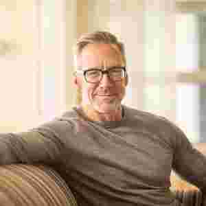 An elderly man is smiling on the couch. White male. Used on home page hero.