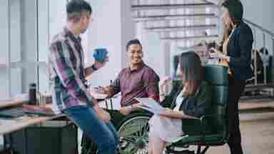 Indian office worker in wheelchair having a cheerful discussion with colleagues in a bright, creative office workspace.