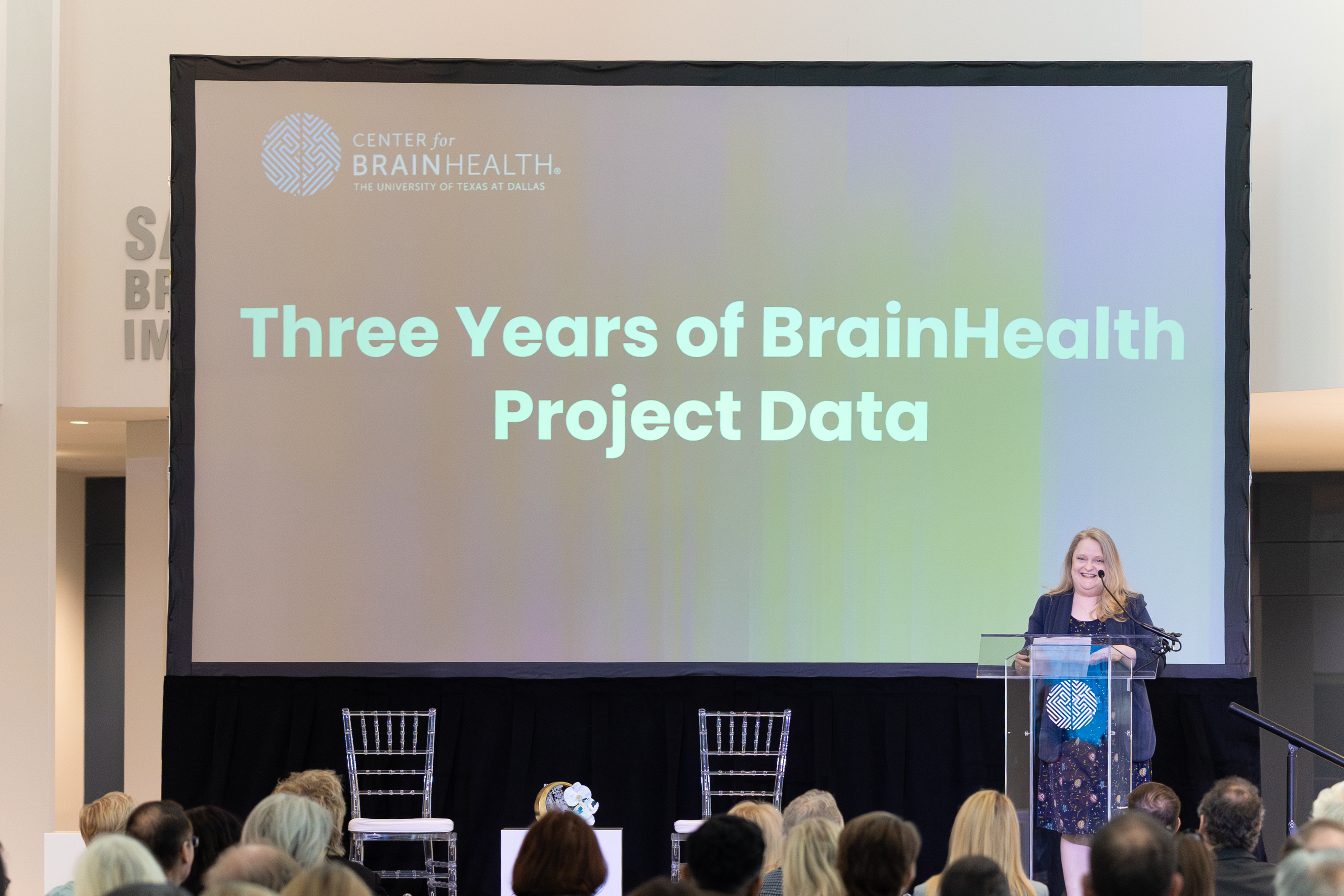 Lori Cook, PhD, CCC-SLP, speaks onstage in front of a large projected screen reading "Three Years of BrainHealth Project Data" to share learnings from the groundbreaking study with the Science Summit audience during BrainHealth Week 2025.