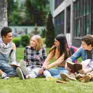 Group of four teens talking while studying at a school campus on the lawn. 
