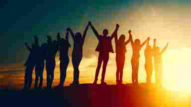 Group of silhouetted people holding hands, arms raised to the sky at sunset. Freedom, friendship and unity concept.