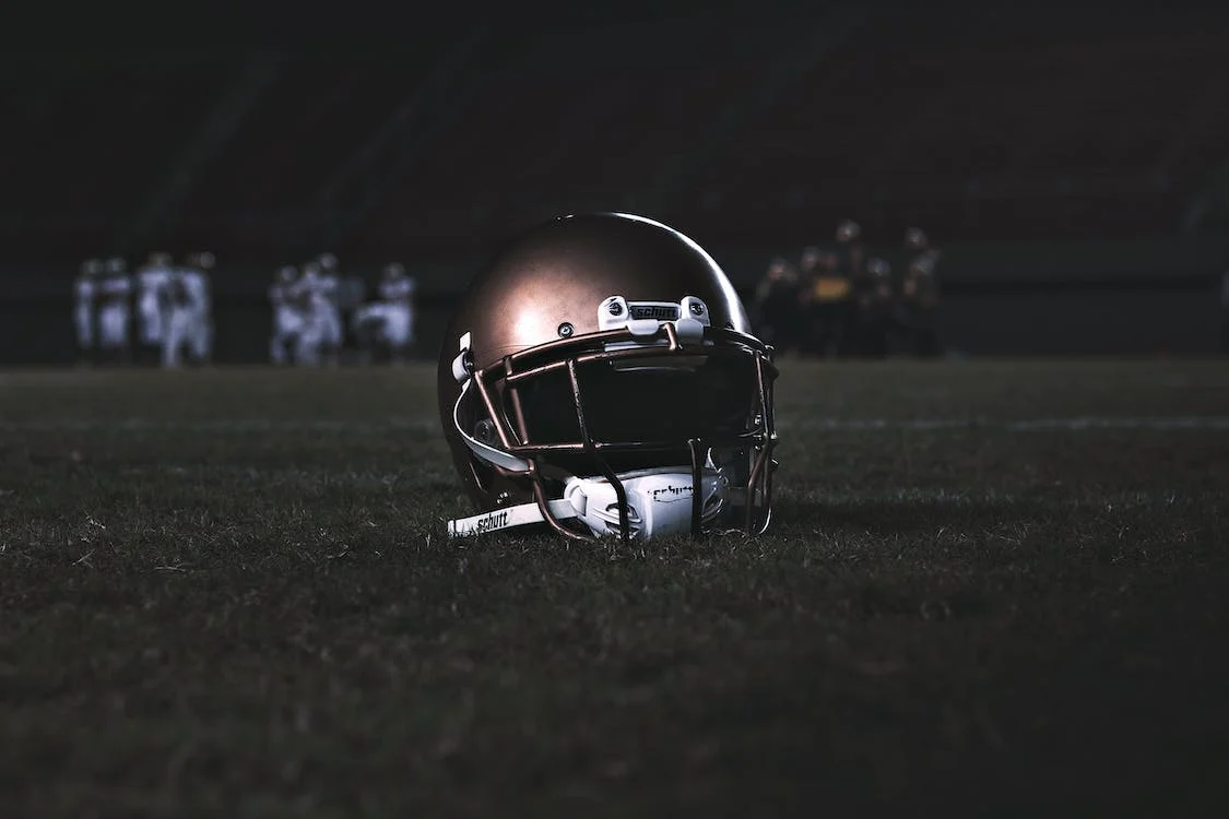 Football helmet on the ground in focus, behind it are players which are in dark lighting and out of focus. 