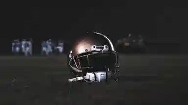 Football helmet on the ground in focus, behind it are players which are in dark lighting and out of focus. 