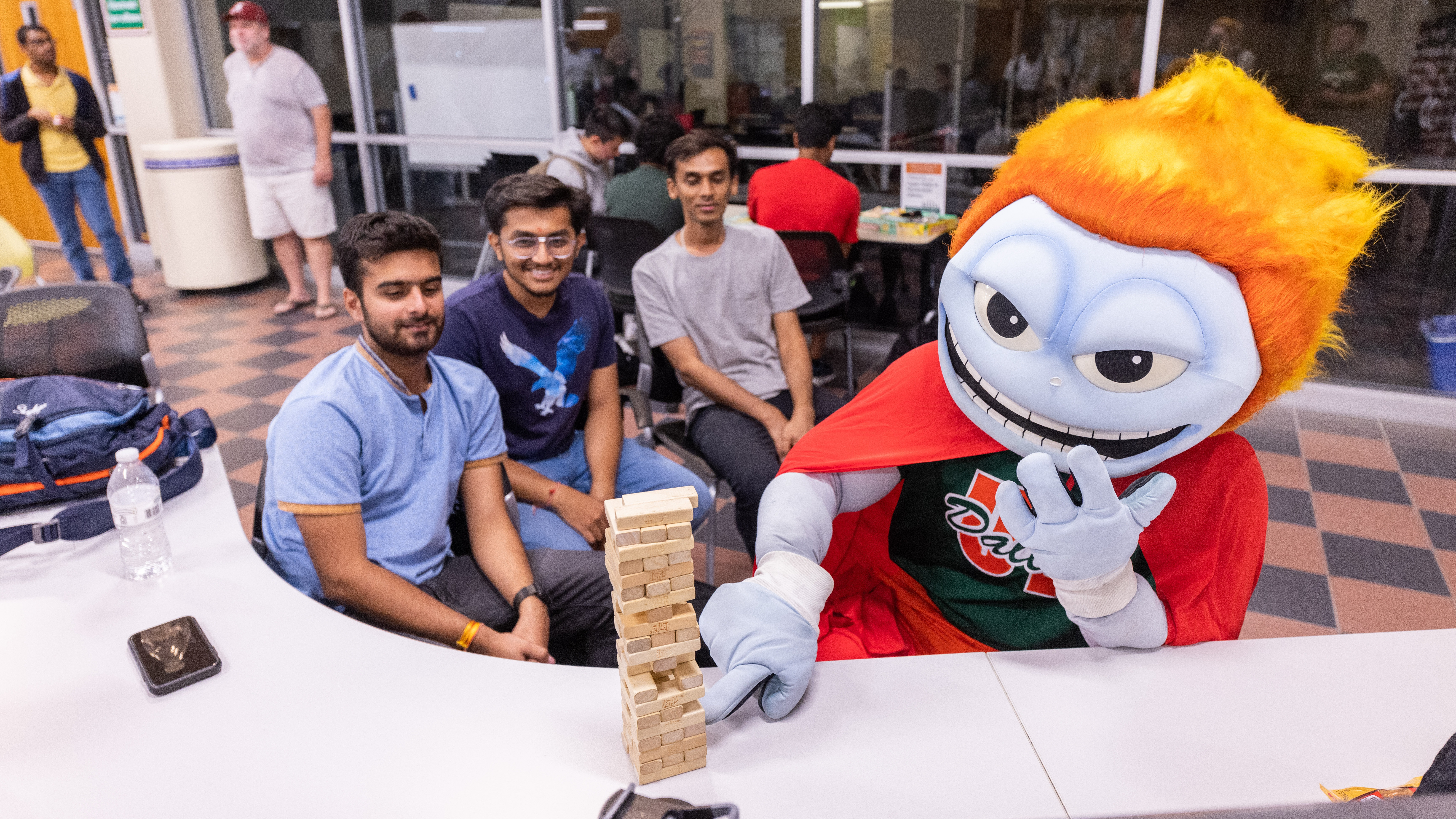 Temoc the Comet, the orange-haired mascot for UT Dallas, plays Jenga with three smiling students at a UTD event.
