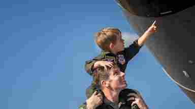 A servicemember shows their young child a real airplane beneath a bright blue sky.
