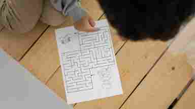 Small child of color solving a labryinth printed on paper sitting on the floor.