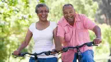 An older, Black married couple happily ride their bikes in a park together.