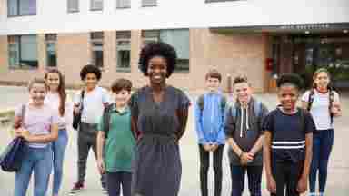 A female teacher wearing a gray dress is standing in front of young students outside the school building. Children