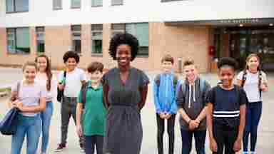 A female teacher wearing a gray dress is standing in front of young students outside the school building. Children