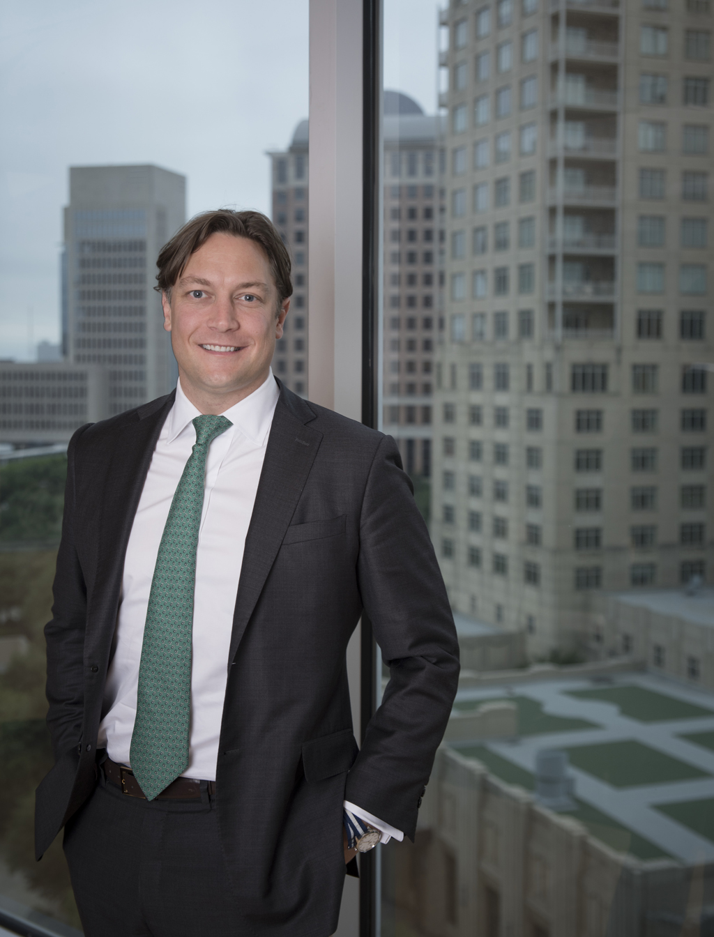 A waist-up shot of Michael Gregory, CIO and Global Head of Highland Alternative Investors. He is wearing a dark blazer and green tie while standing in front of a window with a city view.