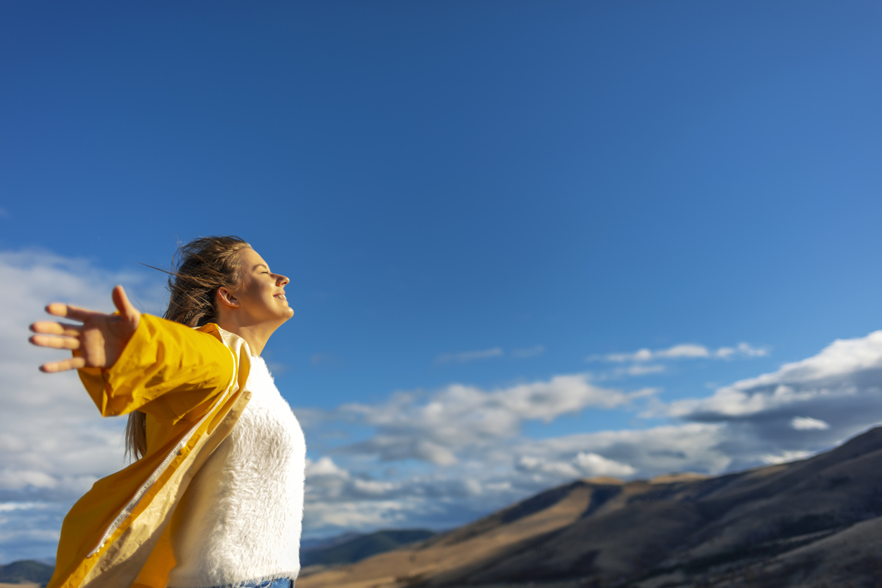 Person in yellow jacket and white sweater with arms outstretched enjoying mountain view under bright blue sky.