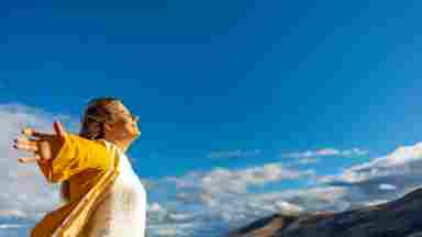 Person in yellow jacket and white sweater with arms outstretched enjoying mountain view under bright blue sky.