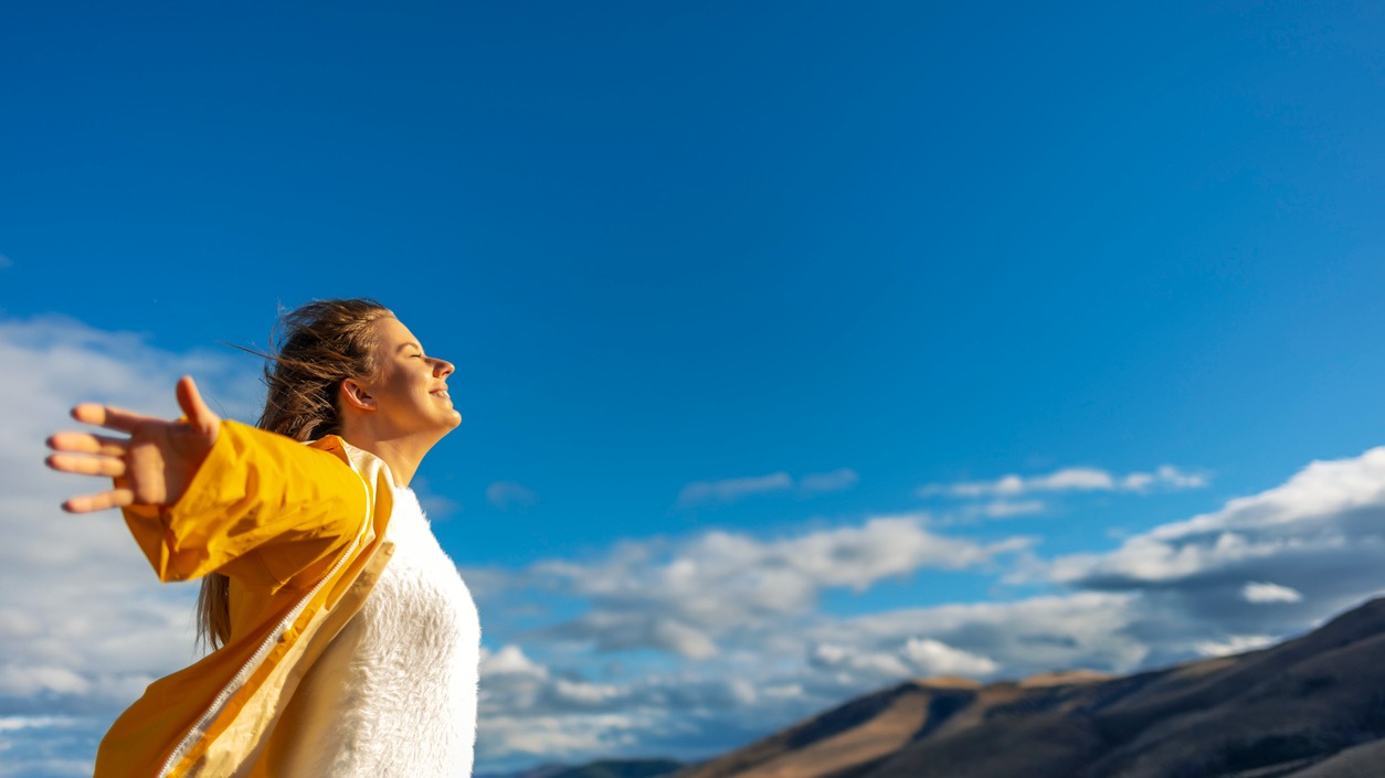 Person in yellow jacket and white sweater with arms outstretched enjoying mountain view under bright blue sky.