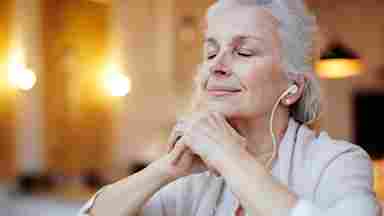 A mature woman with earphones listens to music with a serene expression on her face.