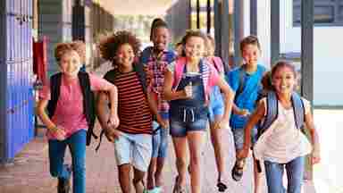 A frontal view of several, diverse elementary school students running in an outdoor corridor at their school.