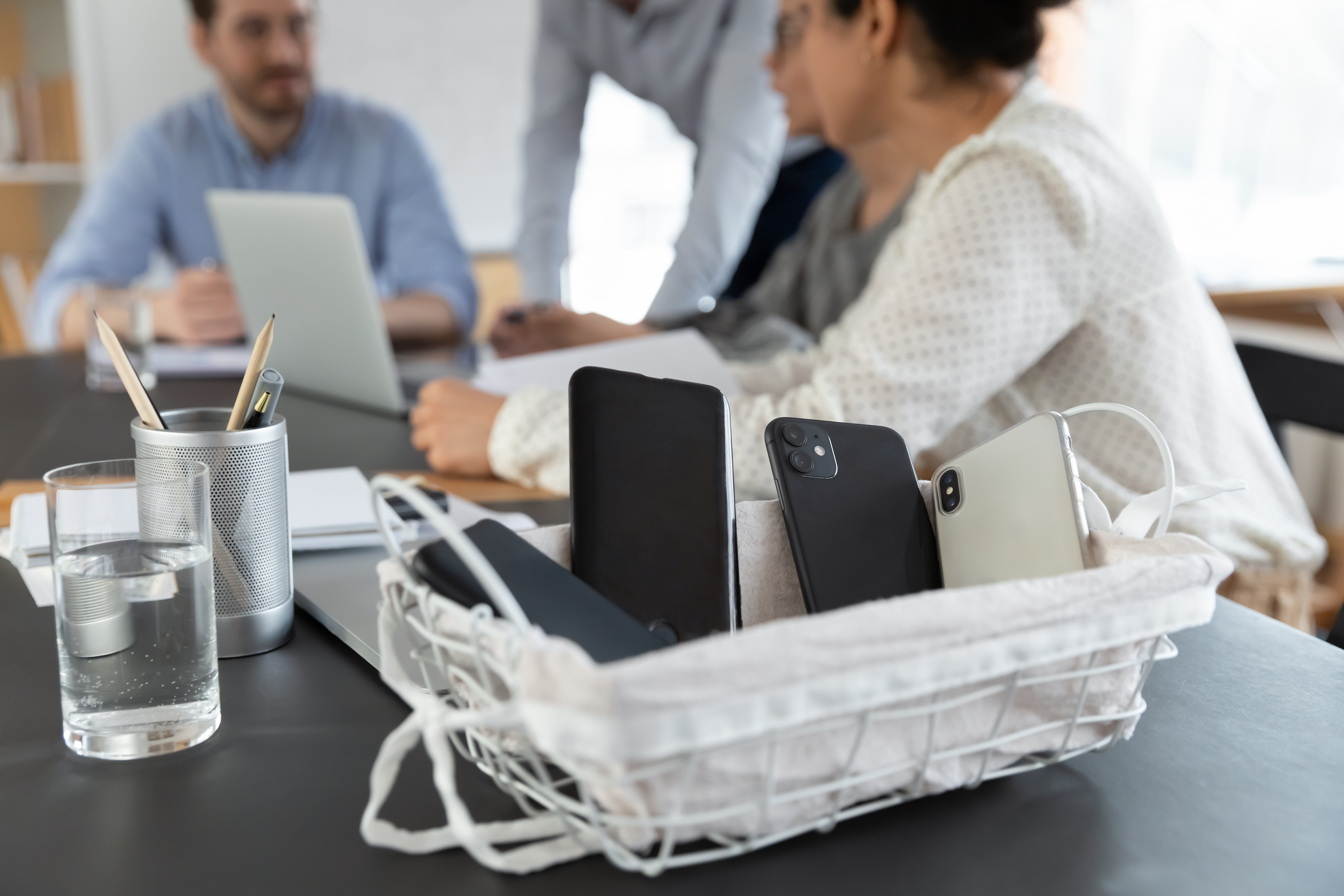 A wire basket holding smartphones and a tablet sits on a meeting table, with colleagues collaborating in the blurred background.
