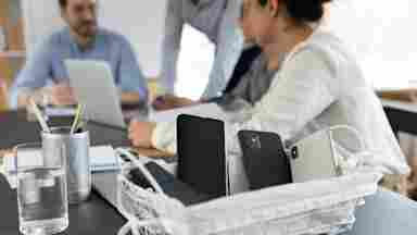 A wire basket holding smartphones and a tablet sits on a meeting table, with colleagues collaborating in the blurred background.
