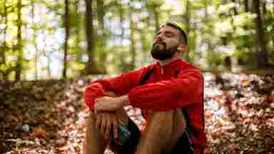 Portrait of a relaxed young man wearing bluetooth headphones sitting on the sunny forest floor.