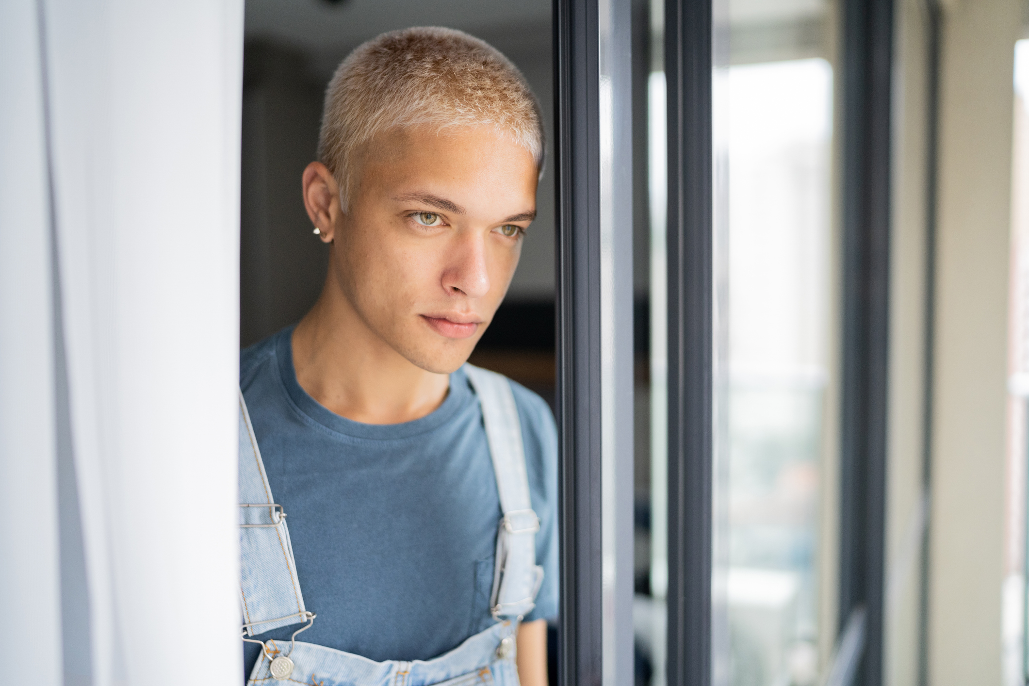 Person with short blonde hair and blue shirt standing by window frame, looking thoughtfully to the side.