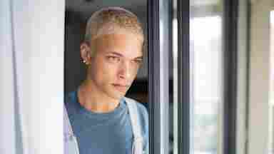 Person with short blonde hair and blue shirt standing by window frame, looking thoughtfully to the side.