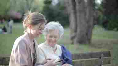 Young woman and smiling elderly woman sitting on park bench while looking at tablet.