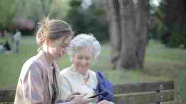 Young woman and smiling elderly woman sitting on park bench while looking at tablet.