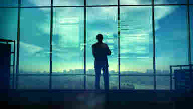 A person stands in silhouette looking over a vast city as computer code reflects against the windows.