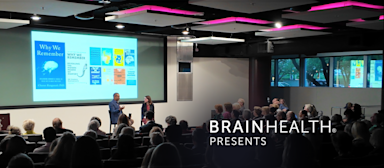 Photograph of neuroscientist Charan Ranganath, PhD, engaged in a fireside chat with Jennifer Zientz during the BrainHealth Presents speaker series, pictured in a crowded auditorium with modern lighting and projected image of publications and media by Dr. Ranganath.