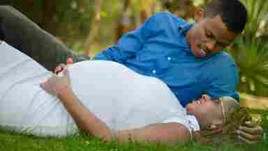 A pregnant woman of color relaxes in the grass at the part with her partner by her side. 