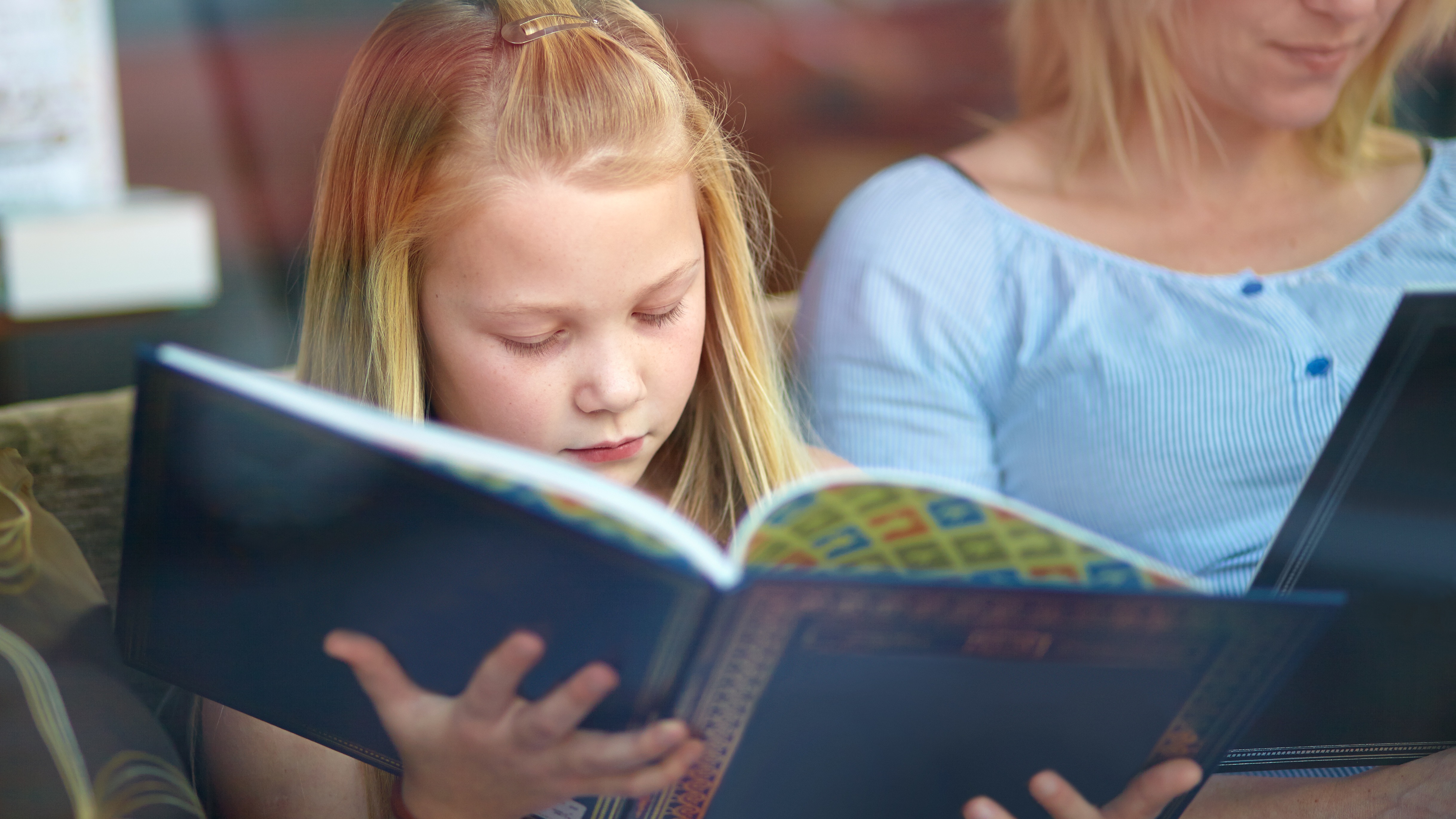 A young blonde girl reads a dark blue book beside a woman in a light blue top also reading, both focused on their books.