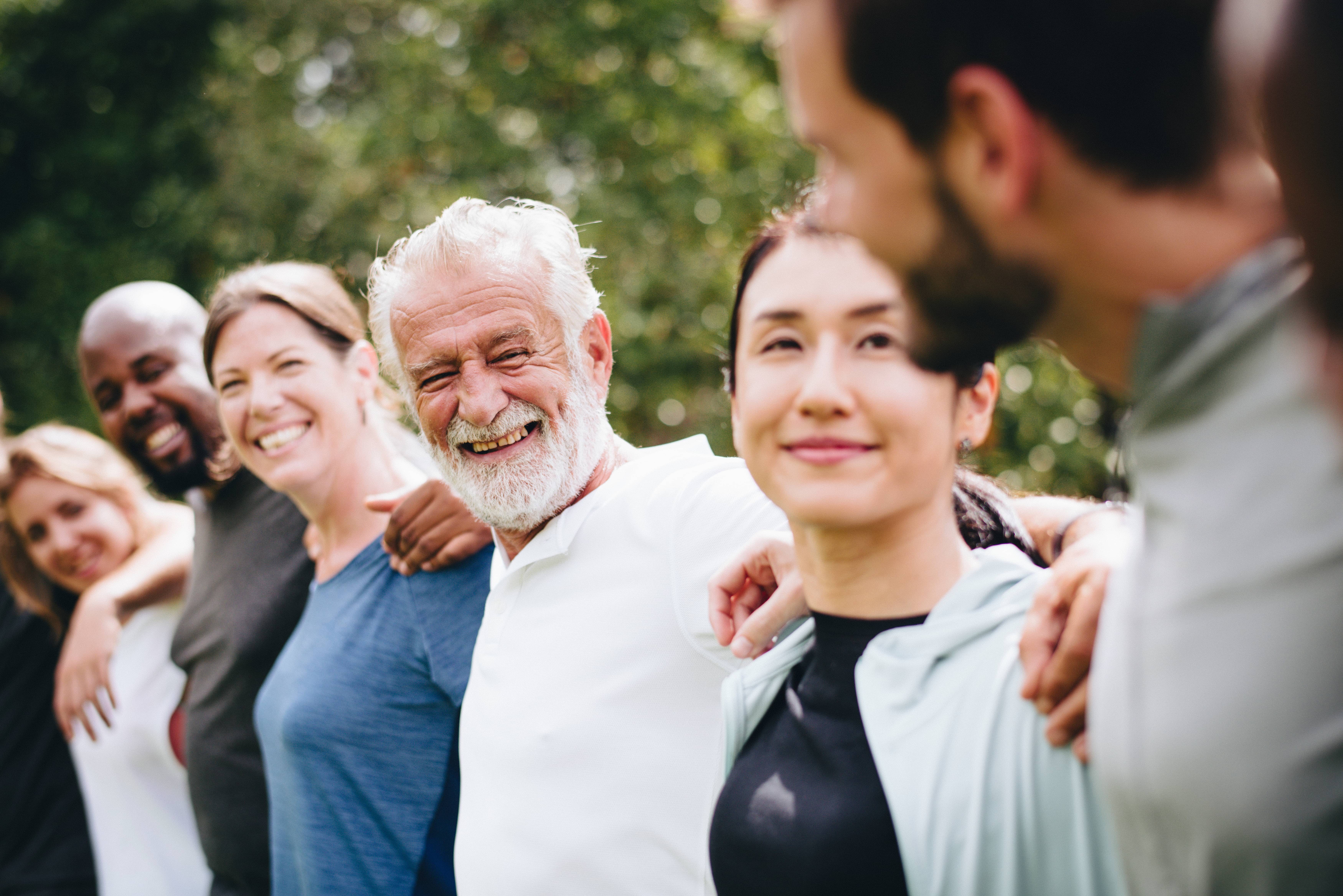 A happy, diverse group of adults standing in a park with their arms around each other's shoulders.