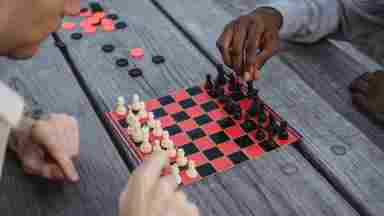 Over the shoulder shot of two people playing chess. The chess board is black and red. A person of color plays the black pieces, and another person plays the white side. 