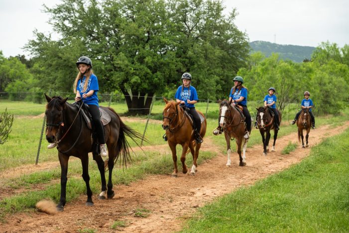 Five children in blue YMCA t-shirts ride horses outdoors along a dirt path
